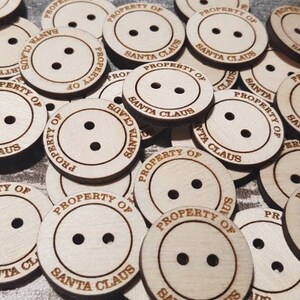 May include: A collection of round, wooden buttons with two holes each. The buttons are light brown and feature the engraved text "PROPERTY OF SANTA CLAUS" around the edge. Ideal for holiday crafts.