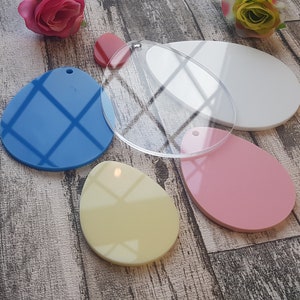 May include: Five acrylic Easter egg shapes in various colours. The eggs are blue, white, clear, pink, and yellow. The eggs are on a wooden surface.