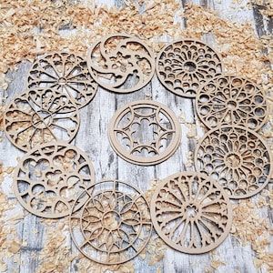 May include: A collection of laser-cut wooden coasters with intricate, openwork designs. The coasters are arranged in a circular pattern on a wooden surface, surrounded by wood shavings. The designs vary, featuring geometric and floral patterns.