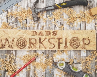 May include: A wooden sign with the words "Dad's Workshop" carved into it. The sign is surrounded by wood shavings and tools, including a hammer, a level, a ruler, and a square.