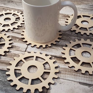 Puede incluir: Cinco posavasos de madera con forma de engranajes, con una taza blanca apoyada en uno de los posavasos. Los posavasos son de madera clara y tienen un acabado natural.