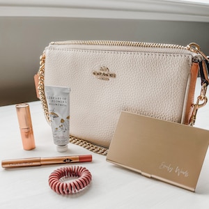 May include: A white leather Coach purse with a gold chain strap, a tube of hand cream labeled "Library of Flowers Willow & Water", a rose gold lipstick, a rose gold lip liner with the text "Charlotte Tilbury", a rose gold hair tie, and a gold metal card holder with the text "Emily Woods" engraved on it.