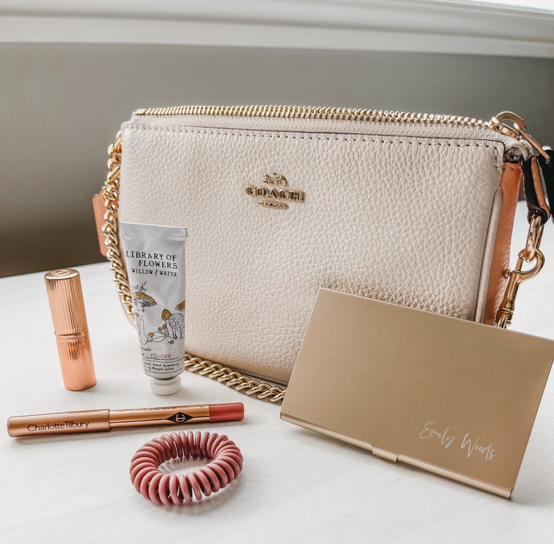 May include: A white leather Coach purse with a gold chain strap, a tube of hand cream labeled "Library of Flowers Willow & Water", a rose gold lipstick, a rose gold lip liner with the text "Charlotte Tilbury", a rose gold hair tie, and a gold metal card holder with the text "Emily Woods" engraved on it.