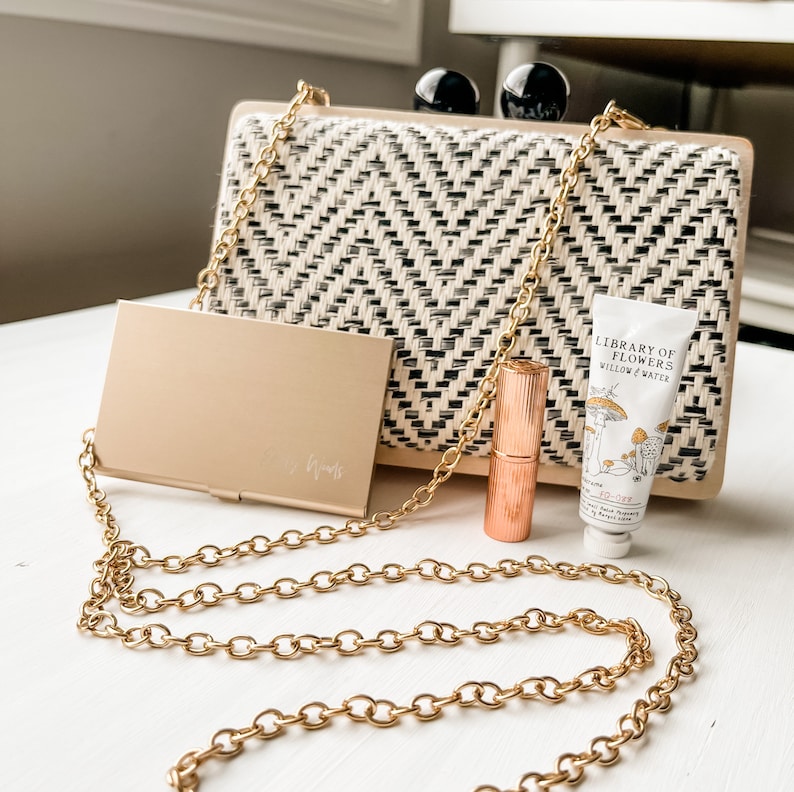 May include: A gold chain purse with a black and white woven pattern. The purse is open and has a gold compact mirror, a rose gold lipstick, and a small tube of hand cream inside. The hand cream tube has a white background with a black and white illustration of mushrooms and the text "Library of Flowers Willow & Water"