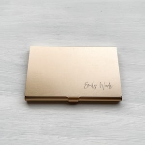 May include: A gold metal business card holder with the name "Emily Woods" engraved on the front.