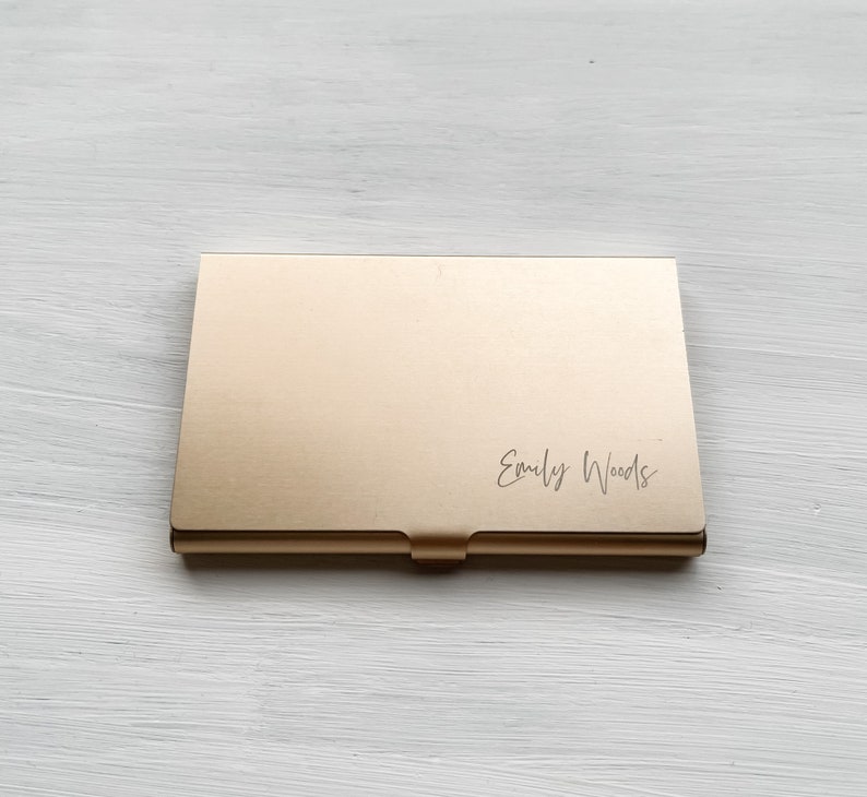 May include: A gold metal business card holder with the name "Emily Woods" engraved on the front.