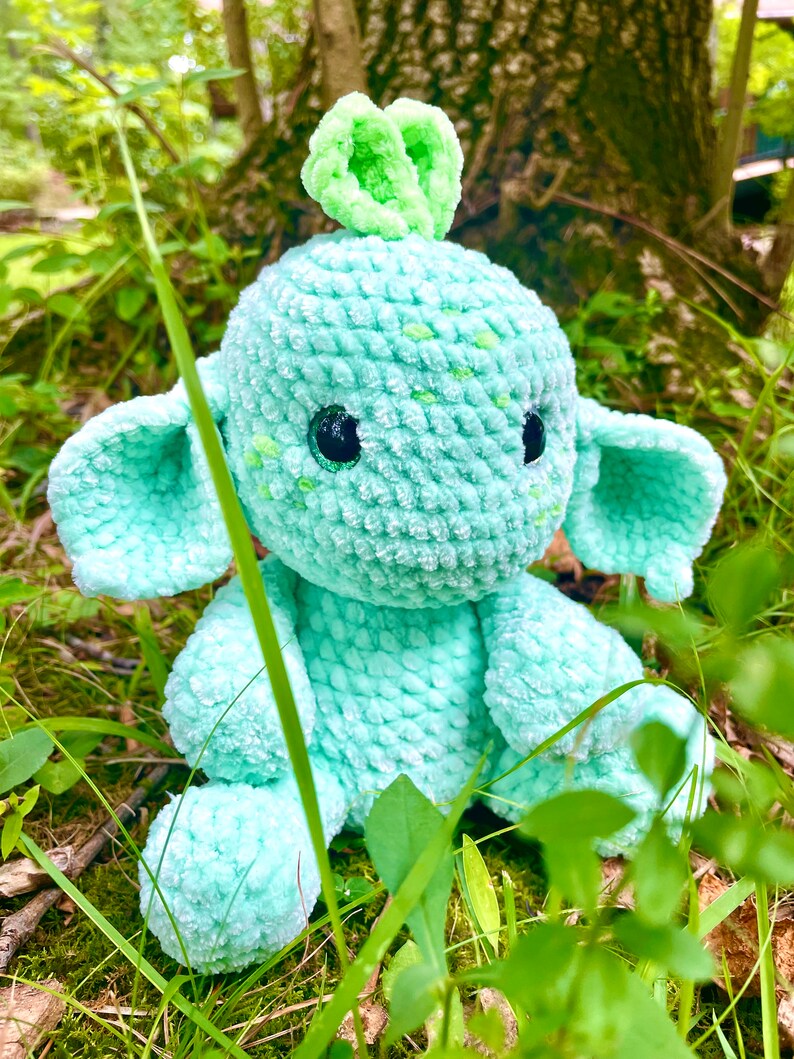 K&ouml;nnte beinhalten: Ein geh&auml;keltes Pl&uuml;schtier in hellgr&uuml;ner Farbe mit gr&uuml;nem Oberteil. Das Spielzeug hat gro&szlig;e Augen und kleine Ohren. Das Spielzeug sitzt in einem Bett aus gr&uuml;nem Gras.