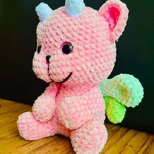 K&ouml;nnte beinhalten: Ein rosa geh&auml;keltes Pl&uuml;schtier-Drache mit einem hellblauen Horn und gr&uuml;nen und hellgr&uuml;nen Fl&uuml;geln. Der Drache hat ein l&auml;chelndes Gesicht mit schwarzen Augen und einer rosa Nase.