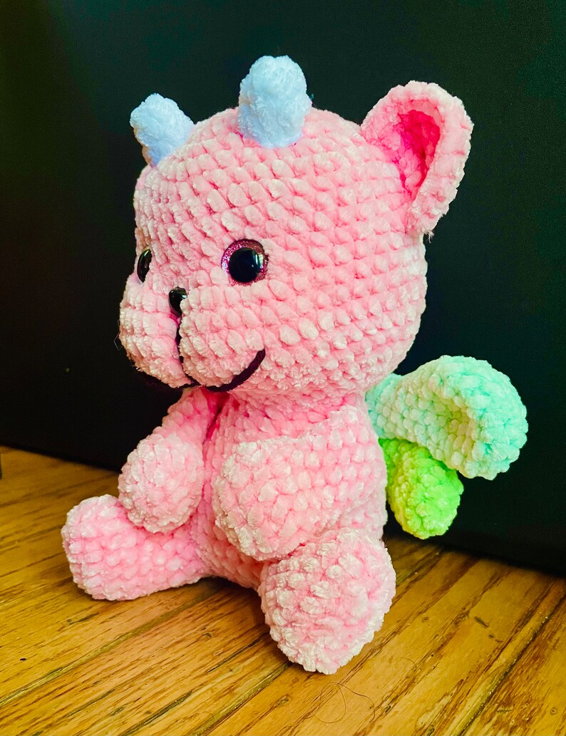 K&ouml;nnte beinhalten: Ein rosa geh&auml;keltes Pl&uuml;schtier-Drache mit einem hellblauen Horn und gr&uuml;nen und hellgr&uuml;nen Fl&uuml;geln. Der Drache hat ein l&auml;chelndes Gesicht mit schwarzen Augen und einer rosa Nase.