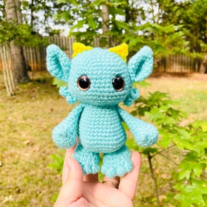 May include: A crocheted blue monster toy with yellow ears and brown eyes. The monster is being held in a hand.