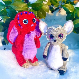 May include: Two crocheted stuffed animals in the snow. One is red with purple fur and large eyes, the other is gray with white fur and large eyes.