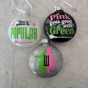 Peut inclure: Trois boules de Noël scintillantes avec des rubans blancs. L'une est blanche avec le texte "You're gunna be POPULAR" en vert et rose. Une autre est noire avec "Pink goes good with Green" en rose et vert. La troisième est argentée avec une silhouette verte et rose.