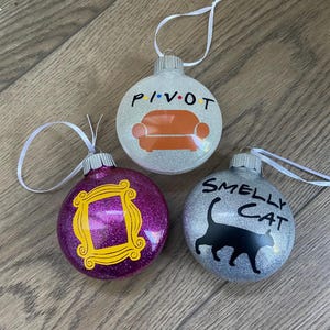 Könnte beinhalten: Drei runde Weihnachtskugeln mit Glitzer. Eine Kugel ist silber mit dem Text "PIVOT" und einer braunen Couch. Eine Kugel ist lila mit einem gelben Rahmen. Eine Kugel ist silber mit dem Text "SMELLY CAT" und einer schwarzen Katze.