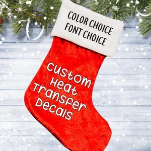 May include: A red Christmas stocking with a white cuff. The text "COLOR CHOICE FONT CHOICE" is printed on the cuff. The text "CUSTOM HEAT TRANSFER DECALS" is printed on the body of the stocking. The background includes Christmas decorations.