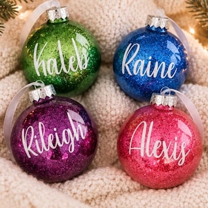 May include: Four round glass Christmas ornaments in green, blue, purple, and pink, each covered in glitter. The ornaments feature white script text reading Kaleb, Raine, Rileigh, and Alexis, and include silver metal caps with white ribbons.