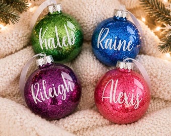 Puede incluir: Cuatro adornos navideños redondos de cristal en verde, azul, morado y rosa, cubiertos de purpurina. Los adornos llevan el texto en blanco con los nombres Kaleb, Raine, Rileigh y Alexis, y tienen tapas de metal plateado con cintas blancas.