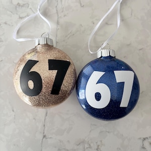 May include: Two round Christmas baubles. One is gold glitter with the number 67 in black. The other is navy blue glitter with the number 67 in white. Both have silver tops and white ribbon hangers.