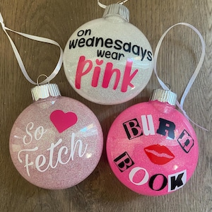 Pu&ograve; includere: Tre palline di Natale glitterate rosa e bianche con citazioni di Mean Girls. La prima pallina dice "On Wednesdays wear pink". La seconda pallina dice "So fetch". La terza pallina dice "Burn Book".