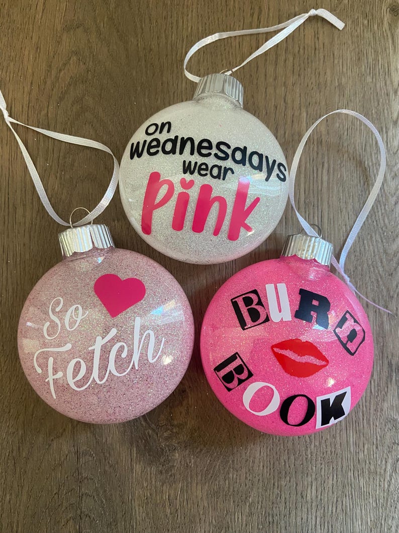 Pu&ograve; includere: Tre palline di Natale glitterate rosa e bianche con citazioni di Mean Girls. La prima pallina dice "On Wednesdays wear pink". La seconda pallina dice "So fetch". La terza pallina dice "Burn Book".
