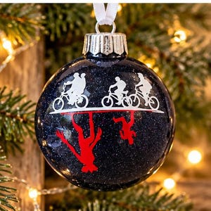 Puede incluir: Un adorno navide&ntilde;o esf&eacute;rico de color azul oscuro con purpurina que presenta un gr&aacute;fico de silueta blanca de tres figuras montando bicicletas sobre una l&iacute;nea horizontal, con un reflejo de silueta roja invertida debajo.
