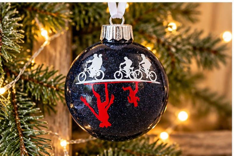 Puede incluir: Un adorno navide&ntilde;o esf&eacute;rico de color azul oscuro con purpurina que presenta un gr&aacute;fico de silueta blanca de tres figuras montando bicicletas sobre una l&iacute;nea horizontal, con un reflejo de silueta roja invertida debajo.