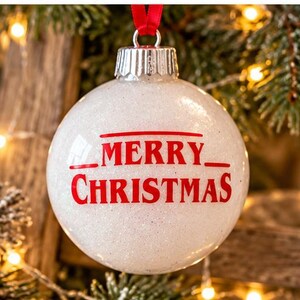 Puede incluir: Un adorno navide&ntilde;o esf&eacute;rico de color blanco con purpurina que presenta el texto rojo MERRY CHRISTMAS impreso en el frente. El adorno tiene una tapa de metal plateado en la parte superior y cuelga de una cinta roja fina.