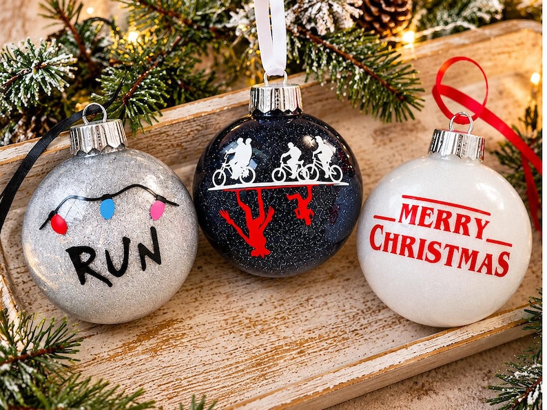 Puede incluir: Tres adornos navide&ntilde;os redondos con cintas para colgar. El de la izquierda es de purpurina plateada con una cadena de luces de colores y la palabra RUN. El central es de purpurina azul oscuro con siluetas blancas y rojas de figuras en bicicleta. El de la derecha es blanco con MERRY CHRISTMAS en rojo.