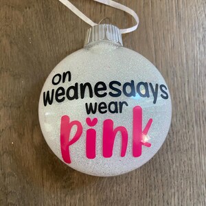 Pu&ograve; includere: Un addobbo natalizio bianco con glitter con il testo "on wednesdays wear pink" in lettere nere e rosa.