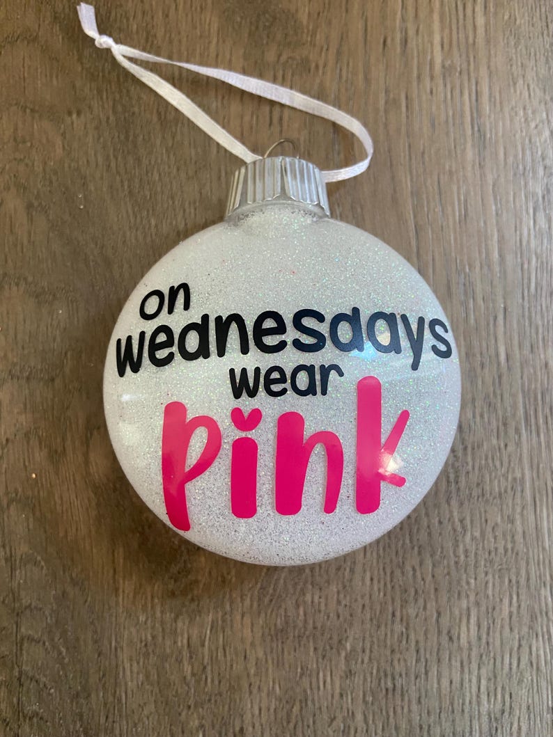 Pu&ograve; includere: Un addobbo natalizio bianco con glitter con il testo "on wednesdays wear pink" in lettere nere e rosa.