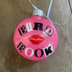 Pu&ograve; includere: Palloncino di Natale rosa con glitter e le parole "BURD BOOK" in lettere bianche e nere. Un'impronta di labbra rosse si trova al centro del palloncino.