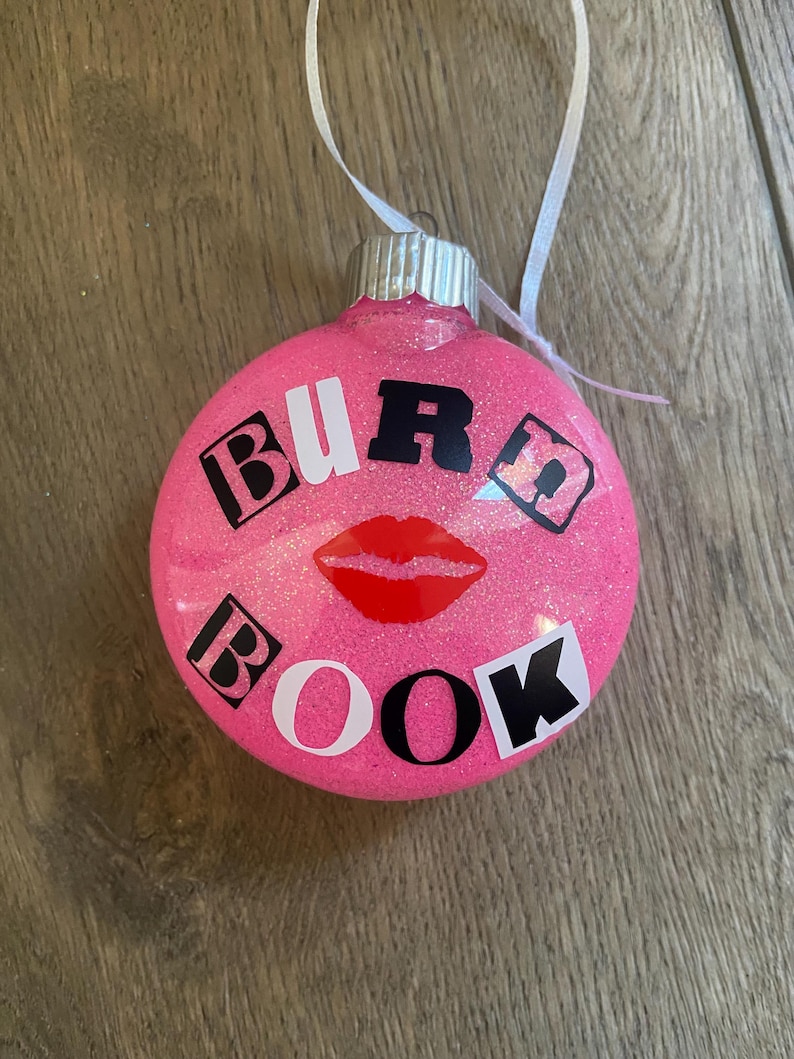 Pu&ograve; includere: Palloncino di Natale rosa con glitter e le parole "BURD BOOK" in lettere bianche e nere. Un'impronta di labbra rosse si trova al centro del palloncino.