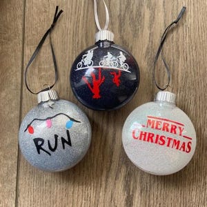 Puede incluir: Tres adornos navide&ntilde;os redondos y brillantes con cintas para colgar. Un adorno plateado muestra una cadena de luces y el texto RUN. Un adorno azul oscuro muestra siluetas blancas de bicicletas. Un adorno blanco dice MERRY CHRISTMAS.