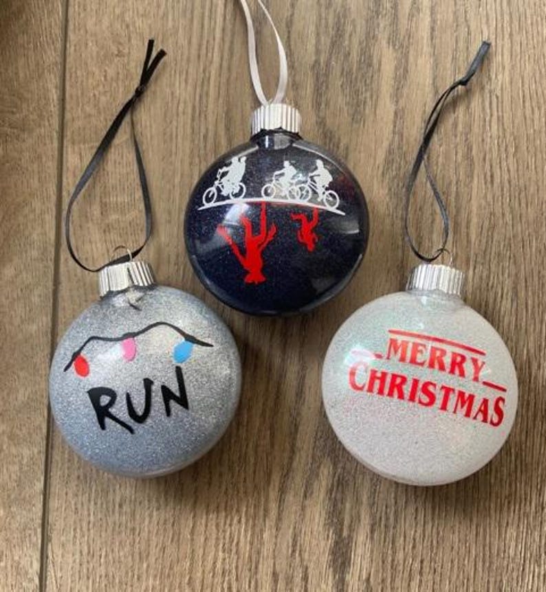 Puede incluir: Tres adornos navide&ntilde;os redondos y brillantes con cintas para colgar. Un adorno plateado muestra una cadena de luces y el texto RUN. Un adorno azul oscuro muestra siluetas blancas de bicicletas. Un adorno blanco dice MERRY CHRISTMAS.