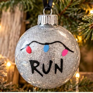 Puede incluir: Un adorno navide&ntilde;o esf&eacute;rico de purpurina plateada con la palabra RUN escrita a mano en negro y una guirnalda de tres peque&ntilde;as bombillas de colores en rojo, azul y rosa. Tiene una tapa de metal plateado y una cinta negra.