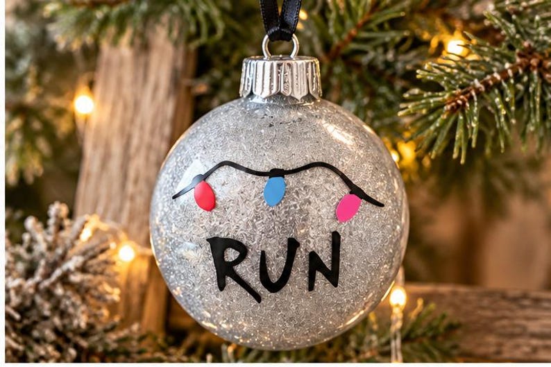 Puede incluir: Un adorno navide&ntilde;o esf&eacute;rico de purpurina plateada con la palabra RUN escrita a mano en negro y una guirnalda de tres peque&ntilde;as bombillas de colores en rojo, azul y rosa. Tiene una tapa de metal plateado y una cinta negra.
