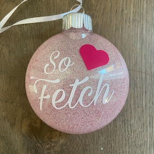 Pu&ograve; includere: Addobbo natalizio rosa glitterato con un nastro bianco e il testo "So fetch" con un cuore rosa.