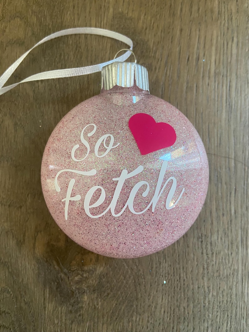 Pu&ograve; includere: Addobbo natalizio rosa glitterato con un nastro bianco e il testo "So fetch" con un cuore rosa.