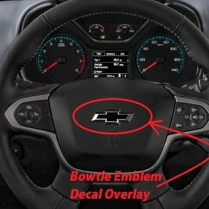 Puede incluir: Un adhesivo negro con el emblema de la pajarita de Chevrolet colocado en el volante de un vehículo. El emblema está rodeado por un óvalo rojo y una flecha roja que señala hacia él. El texto Bowtie Emblem Decal Overlay aparece en rojo.