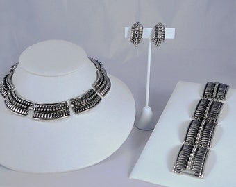 May include: A silver necklace, bracelet, and earrings set with a geometric design. The jewelry is made of linked, curved, and beaded pieces.