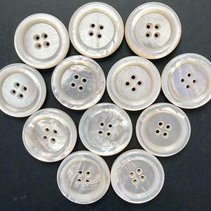 May include: A collection of eleven round, off-white buttons with four holes each. The buttons have a pearlescent sheen and a slightly raised center. The buttons are arranged in a circular pattern against a dark background.