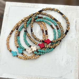 May include: A collection of beaded bracelets in various colors, including turquoise, brown, and cream. Each bracelet features a cross-shaped charm in red, white, or turquoise. The bracelets are displayed on a white surface.
