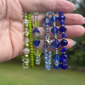 Puede incluir: Una colección de pulseras de cuentas en tonos verdes y azules. Las pulseras presentan varias formas y tamaños de cuentas, incluyendo diseños facetados, redondos y florales. Algunas pulseras tienen detalles y dijes plateados.