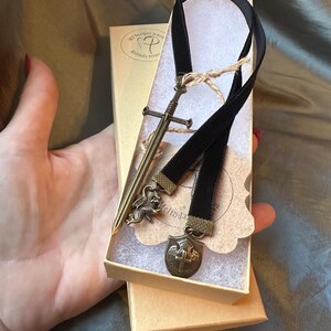 Op de afbeelding: Een zwarte fluwelen bladwijzer met een zwaard-, draak- en schildbedel. De bladwijzer is verpakt in een kleine bruine doos met de tekst "My Designs Jewelry Melinda Jerrell" op de deksel.