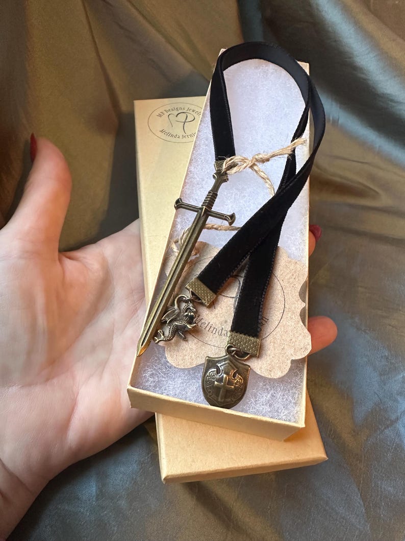 Op de afbeelding: Een zwarte fluwelen bladwijzer met een zwaard-, draak- en schildbedel. De bladwijzer is verpakt in een kleine bruine doos met de tekst "My Designs Jewelry Melinda Jerrell" op de deksel.