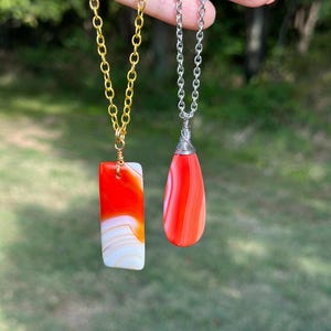 Puede incluir: Dos collares con colgantes de piedra de ágata roja y blanca. Un colgante es de forma rectangular y el otro es de forma de lágrima. Los collares están en una cadena dorada y plateada.