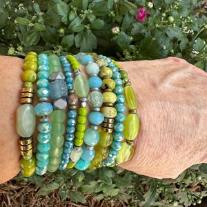 May include: A stack of bracelets in shades of green and blue. The bracelets are made of various materials, including glass beads, metal beads, and stones.