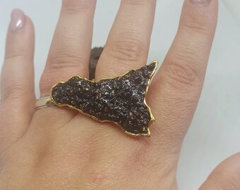 May include: A gold ring with a black, textured, irregular shape. The ring is worn on a finger.