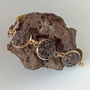 Può includere: Bracciale dorato con pietre ovali, scure e macchiate, incastonate in castoni dorati. Il bracciale è esposto su una superficie di pietra grezza, marrone, che mette in risalto la consistenza e il contrasto cromatico del gioiello. Un accessorio delicato ed elegante.
