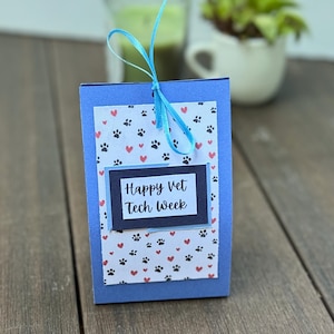 May include: A blue card with a white background and a black rectangle with the text "Happy Vet Tech Week". The card has a blue ribbon tied around it and a pattern of paw prints and hearts.