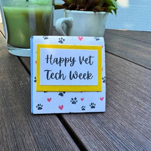 May include: A small card with a yellow border and a white background with black paw prints and pink hearts. The card says "Happy Vet Tech Week" in black text.
